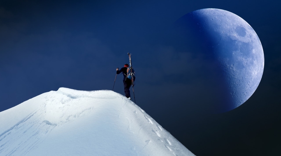 Muž s batohom na zasneženom kopci a mesiac na oblohe